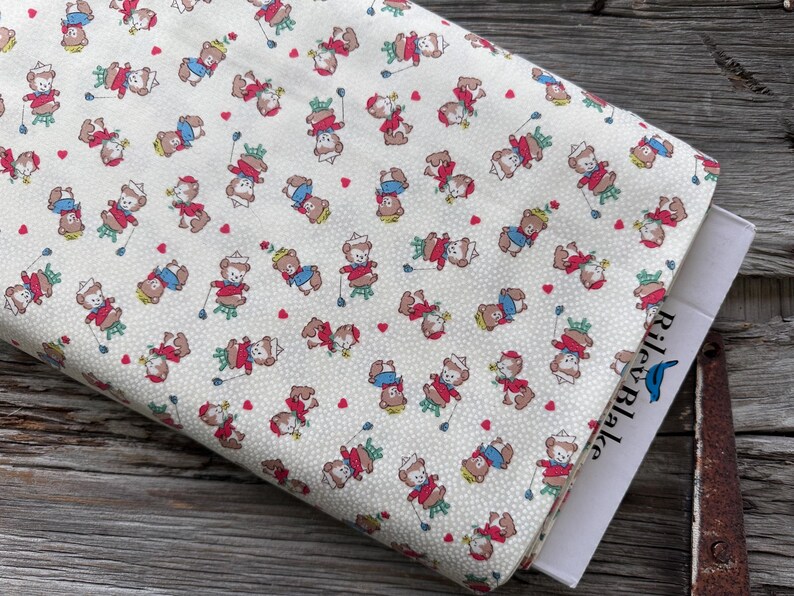 May include: Cream-colored fabric with a repeating pattern of cartoon bears in red, blue, and white outfits, holding golf clubs. Small red hearts are scattered throughout the design. A book with the text "Riley Blake" is visible.