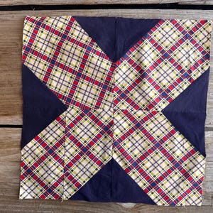 May include: Square fabric block with a navy blue background and four plaid sections in red, green, and white. The plaid sections are arranged in a symmetrical pattern, creating an X shape.