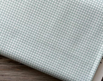 May include: A folded piece of fabric featuring a small-scale gingham pattern in light sage green and white. The textile displays a consistent grid design across the surface, with a white paper tag visible at the bottom right corner.