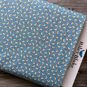 May include: A folded fabric with a repeating pattern of small, abstract shapes in cream and red on a light blue background. The fabric is partially covering a white label with the text "Riley Blake Designs".