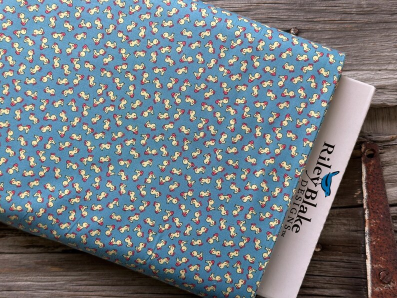 May include: A folded fabric with a repeating pattern of small, abstract shapes in cream and red on a light blue background. The fabric is partially covering a white label with the text "Riley Blake Designs".