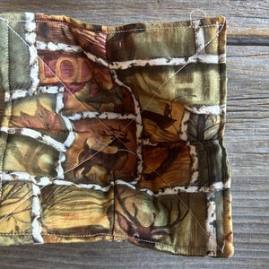 Puede incluir: Posavasos cuadrado de tela acolchada con un estampado de temática natural. El diseño presenta un patchwork de cuadrados de tela verdes, marrones y beiges con imágenes de hojas y ciervos. Las costuras blancas delinean los cuadrados.