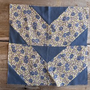 May include: A square patchwork quilt block with a floral pattern in blue, green, and cream, set against a navy blue background. The design features a central navy blue cross with angled navy blue corners.