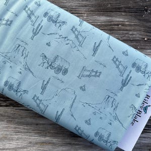 May include: A light blue fabric with a repeating pattern of a wagon pulled by horses, cacti, and fences. The fabric is folded and a white tag with the text "Riley Blake" is visible.