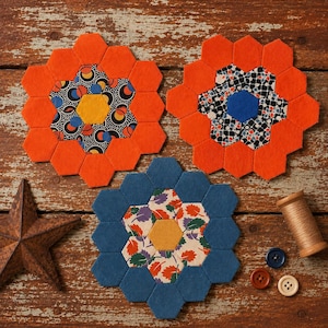 May include: Three fabric hexagon flower motifs made from patchwork pieces. Two motifs feature bright orange outer petals, one with a yellow center and black patterned fabric, the other with a blue center and floral print. One motif has blue petals.
