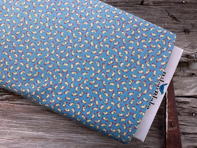 May include: A folded piece of blue fabric with a repeating pattern of small, abstract figures in cream and red. The fabric is resting on a wooden surface, with a white paper partially visible. The paper has the text "pixelable" and a blue bird logo.