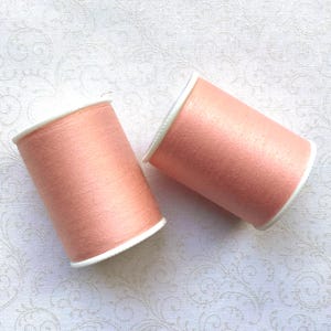 May include: Two spools of peach-colored thread with white rims are displayed against a white patterned background. The thread appears to be tightly wound, and the spools are cylindrical in shape. The image is well-lit, highlighting the texture of the thread.