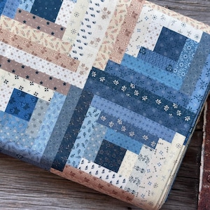 Op de afbeelding: Een opgevouwen quilt met een blokhutontwerp. De quilt heeft verschillende tinten blauw, crème en bruin. De stofpatronen omvatten bloemen-, ster- en geometrische ontwerpen. De quilt is opgevouwen en toont de gelaagde constructie en gedetailleerde stiksels.
