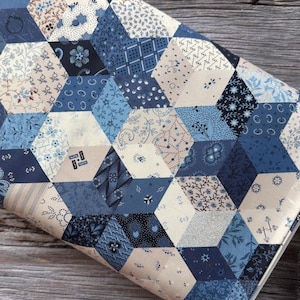 Puede incluir: Tela doblada con un patrón geométrico de formas hexagonales azules y crema. Cada hexágono presenta un diseño floral o estampado diferente, creando un efecto de patchwork. La tela parece ser de algodón o un material similar.