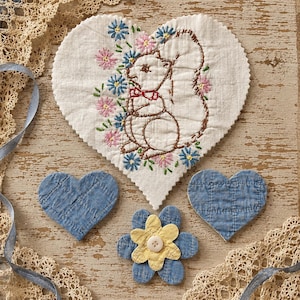 May include: A large heart-shaped white fabric piece featuring brown embroidered squirrel and colorful floral accents, accompanied by two smaller blue quilted fabric hearts and a blue and yellow fabric flower with a central white button.