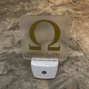 K&ouml;nnte beinhalten: Ein wei&szlig;es Nachtlicht mit einer quadratischen, durchsichtigen Plastikplatte, auf der ein goldener griechischer Buchstabe Omega und der Text "Omega Psi Phi" zu sehen sind.