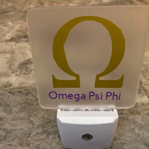 K&ouml;nnte beinhalten: Ein wei&szlig;es Nachtlicht mit einer mattierten quadratischen Platte, auf der ein goldener griechischer Buchstabe Omega und der Text "Omega Psi Phi" zu sehen sind.