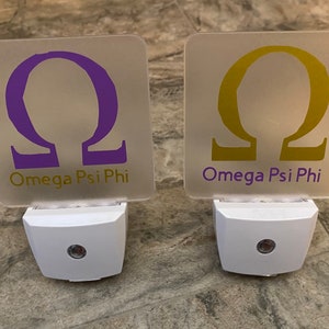 K&ouml;nnte beinhalten: Zwei wei&szlig;e Nachtlichter mit mattierten Glasplatten. Die Platten zeigen den griechischen Buchstaben Omega in Lila und Gold, mit dem Text "Omega Psi Phi" darunter.
