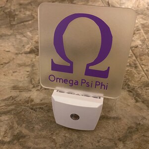 K&ouml;nnte beinhalten: Ein wei&szlig;es Nachtlicht mit einer durchsichtigen quadratischen Plakette mit einem violetten griechischen Buchstaben Omega und dem Text "Omega Psi Phi".