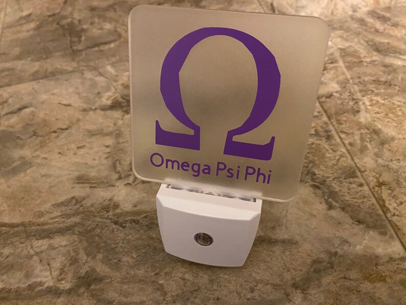 K&ouml;nnte beinhalten: Ein wei&szlig;es Nachtlicht mit einer durchsichtigen quadratischen Plakette mit einem violetten griechischen Buchstaben Omega und dem Text "Omega Psi Phi".