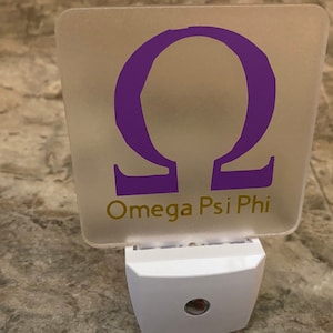 K&ouml;nnte beinhalten: Ein wei&szlig;es Nachtlicht mit einer mattierten Glasscheibe, auf der ein violetter griechischer Buchstabe Omega und der Text "Omega Psi Phi" in goldenen Buchstaben zu sehen sind.