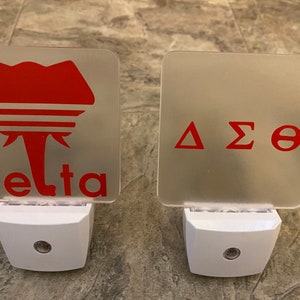 May include: Two white night lights with frosted glass covers. The left night light has a red elephant graphic with the word "delta" below it. The right night light has the Greek letters "ΔΣΘ" in red.
