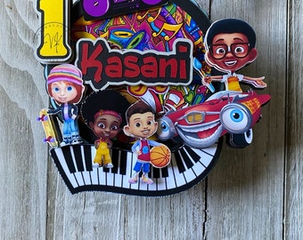 May include: A colorful cake topper featuring a cartoon character with a red car, a piano, a boombox, and the name "Kasani".