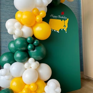 May include: A festive balloon garland in white, yellow, and green, with a green backdrop featuring a golf course map and the words "Happy Birthday." The balloons are arranged in clusters and arches, creating a celebratory atmosphere.