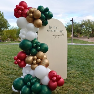 May include: A beige archway backdrop with the text "Tis the season to get engaged!" decorated with a garland of green, white, gold, and red balloons.