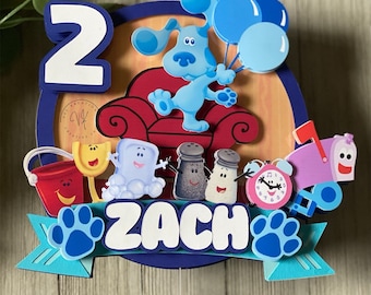 May include: A colorful cake topper featuring Blue's Clues characters, a red couch, and blue balloons. The number "2" is displayed, along with the name "ZACH" on a banner, and paw prints. The topper is designed for a birthday celebration.