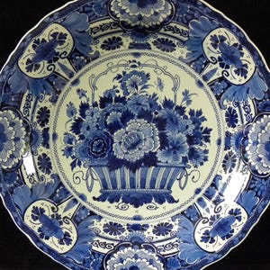 Può includere: Un piatto rotondo in ceramica con bordo smerlato caratterizzato da un motivo floreale blu e bianco. Il centro mostra un cesto pieno di fiori vari, mentre il bordo è decorato con intricati motivi di fogliame e fiori blu.