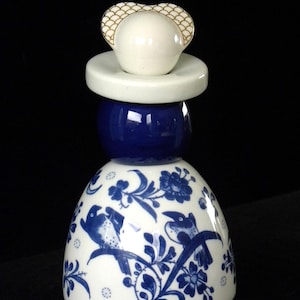 May include: A white ceramic bottle stopper with a blue and white floral design and two birds. The stopper has a white cap with a gold heart design.