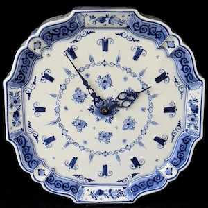 Könnte beinhalten: Eine dekorative Keramikuhr mit weißem Hintergrund und blauen Blumen- und Scrollwork-Designs. Das Zifferblatt hat blaue Zeiger und ein zentrales Blumenmotiv. Die Uhr hat einen gewellten Rand und einen Durchmesser von etwa 25 cm.