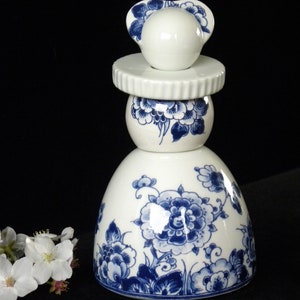 May include: A white ceramic bottle with a blue and white floral design. The bottle is shaped like a woman wearing a hat.