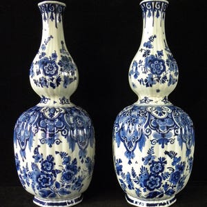 May include: Two matching white and blue ceramic vases. The vases have a bulbous base, a mid-section, and a slender neck. The blue floral design covers the entire surface of the vases. The vases are displayed against a black background.