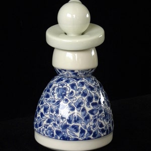 May include: A white ceramic bottle stopper with a blue and white floral design. The stopper is shaped like a woman wearing a dress.