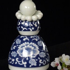 May include: A decorative porcelain bottle stopper with a blue and white floral design. The stopper has a bulbous base, a mid-section with a floral pattern, and a white top with a spherical finial. The stopper is approximately 15 cm tall.