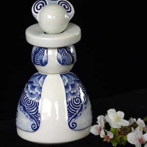 May include: A white ceramic figurine of a woman wearing a blue and white patterned dress. The figurine has a round head with two blue and white swirls on top. The figurine is standing on a black surface.