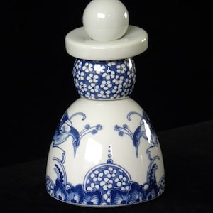 May include: A white ceramic bottle stopper with a blue and white floral design. The stopper is shaped like a woman wearing a traditional Dutch dress.