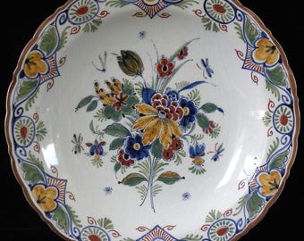Könnte beinhalten: Ein runder Keramikteller mit gewelltem Rand und einem zentralen, handgemalten Blumenstrauß in Blau-, Gelb-, Grün- und Rottönen. Der Rand ist mit wiederkehrenden Blumenmotiven und einem braunen Saum auf weißem Grund verziert.