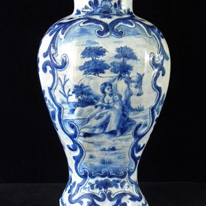 Könnte beinhalten: Eine weiß-blaue Keramikvase mit einer ländlichen Szene. Die Vase hat ein blau-weißes Design mit einer Frau, einer Kuh und Bäumen. Die Vase hat einen breiten Fuß und einen schmalen Hals, mit einer Höhe von etwa 25 cm.