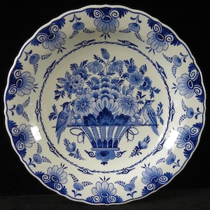 May include: A round ceramic plate with a scalloped edge, featuring a white base and a blue hand-painted design. The center displays a basket filled with flowers and two birds perched on the sides. The rim is decorated with floral motifs.