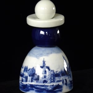 May include: A blue and white ceramic bottle stopper shaped like a Dutch woman wearing a traditional white cap. The bottle stopper features a blue and white Delftware design of a Dutch town.