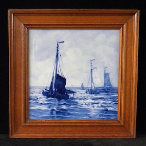 May include: A framed nautical painting depicts sailboats on a wavy sea under a cloudy sky. The artwork is primarily blue and white, with a wooden frame. The painting's style is reminiscent of Delftware.