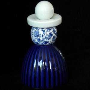 May include: A blue and white ceramic doll figurine with a white hat and a blue and white patterned skirt. The doll is standing on a black surface.