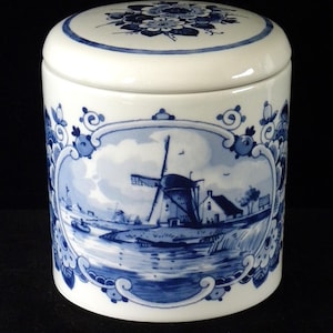 May include: A white ceramic jar with a blue and white floral design and a scene of a windmill in a Dutch landscape. The jar has a lid.