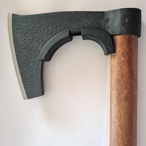 Könnte beinhalten: Axt mit einem dunkelgrauen Kopf und einem Holzgriff. Der Axtkopf hat eine strukturierte Oberfläche mit kleinen Löchern und einem gebogenen Schutz. Der Griff ist aus poliertem Holz. Die Axt ist zum Hacken und Spalten von Holz bestimmt.