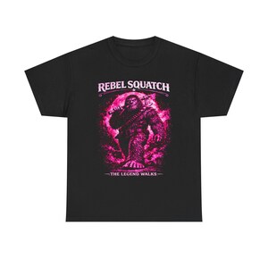 May include: Black t-shirt featuring a pink graphic of a Bigfoot-like creature holding a weapon. The text "REBEL SQUATCH" is arched above the image, with "-THE LEGEND WALKS-" below. The design has a distressed, vintage look.
