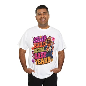 Pode incluir: Camiseta branca com um gráfico colorido de um homem apontando, com as palavras "SHIP BUILD REPEAT BABY YEAH!!" em uma fonte retrô. O design inclui flores e um arco-íris.