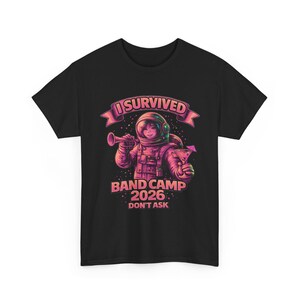 Puede incluir: Camiseta negra con un gráfico colorido de un astronauta tocando la trompeta y sosteniendo un cóctel. El texto dice "I Survived Band Camp 2026 Don't Ask" en rosa y blanco.