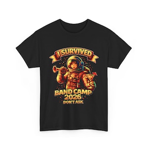 Puede incluir: Camiseta negra con un gráfico colorido de un astronauta tocando la trompeta y sosteniendo un cóctel. El texto dice "I Survived Band Camp 2026 Don't Ask". El astronauta lleva un traje espacial.