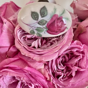 May include: A white ceramic knob with a pink rose and green leaves design. The knob is sitting on top of a bouquet of pink roses.