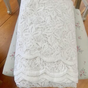 May include: A folded roll of white lace fabric with a detailed floral pattern. The lace has a scalloped edge and is resting on a floral patterned cushion. The fabric is likely used for sewing or crafting projects.