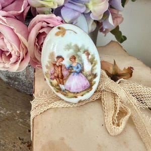 May include: A white porcelain egg-shaped trinket box with a painted scene of a couple in period clothing. The box has a gold leaf accent and is resting on a stack of aged paper with a cream-coloured lace ribbon. Pink and purple flowers are in the background.