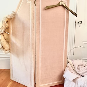 May include: A folding room divider upholstered in pale pink velvet with white trim detailing. A gold-toned wooden clothes hanger with a floral decal hangs from the top edge of the divider. The divider features a curved top panel on one side.